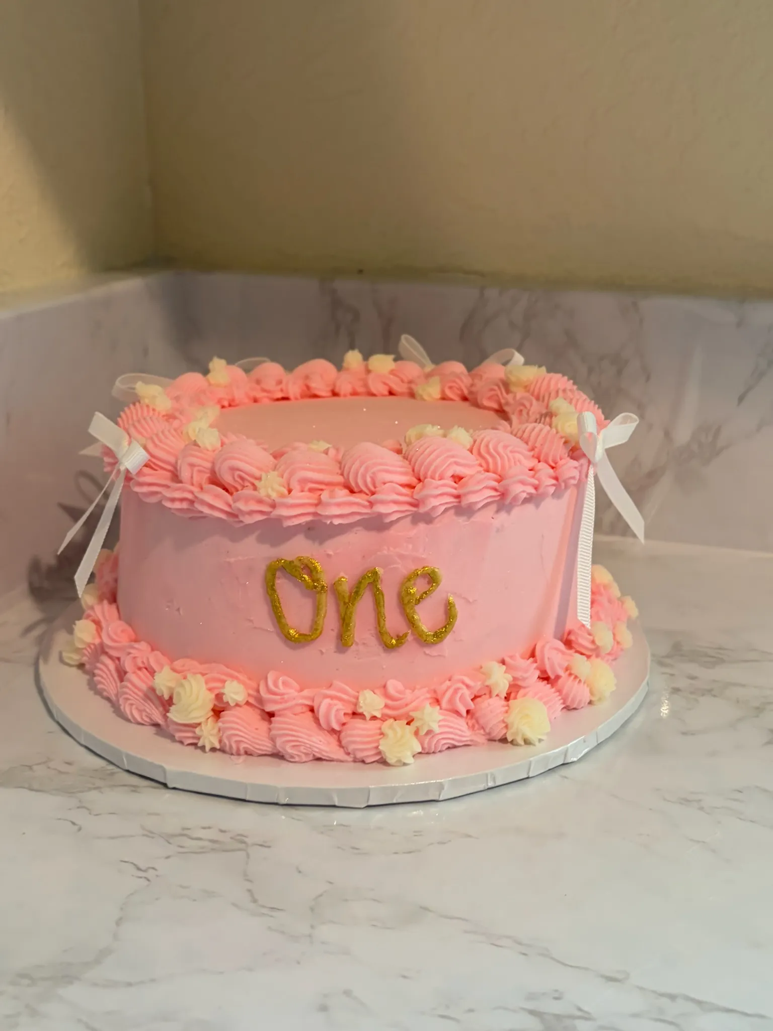 A pink buttercream first-birthday cake with ribbon bows and a gold 'One' topper