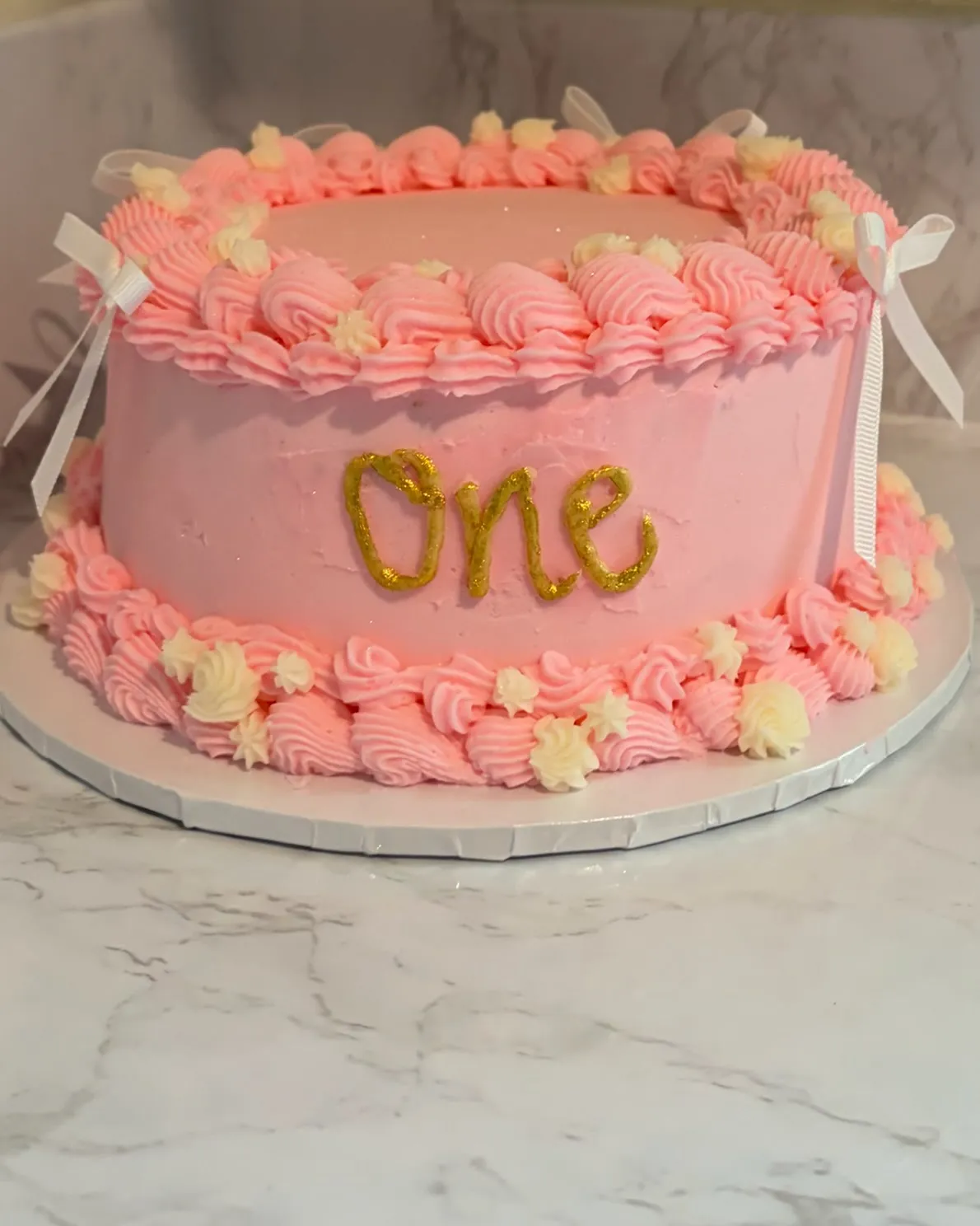 A pink buttercream first-birthday cake piped with shell borders, ribbon bows and a gold 'One' topper